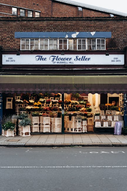 The exterior of a flower shop named 'The Flower Seller of Muswell Hill', with a large glass display window showcasing an array of colorful fresh flowers arranged on multiple tiered shelving units, some in vases and some in pots. The shop's signage is visible above the window, with the name and contact details. The street in front is paved, with a white line marking the parking or traffic zone. The building's facade includes a brick upper section and a corrugated canopy over the entrance, which appears to be in a commercial area. The scene is evenly lit, with natural daylight highlighting the vibrant flower displays. This setting relates to domestic or commercial surface cleaning, and suggests the importance of maintaining tidy, hygienic storefronts, aligning with the services offered by Oven Cleaning Haringey in their professional cleaning and sanitation offerings.