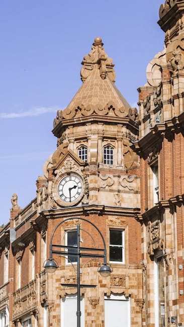 Close-up view of a historic sandstone building with intricate architectural details, including a domed turret topped with a finial, a clock face showing the time as approximately 4:15, and large arched windows. The structure is illuminated by natural daylight, highlighting the warm tones and detailed carvings on the facade. A black street lamp with two round lights is visible in the foreground, mounted on a tall pole, situated on a city street. The background features a clear blue sky, contributing to the well-lit scene. This image emphasizes the architectural beauty of the building's surface and their clean, well-maintained appearance, consistent with expert surface cleaning and preservation practices, as provided by Oven Cleaning Haringey for residential and commercial properties, including deep cleaning and sanitisation services in the Haringey area.