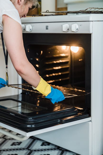 Close-up of a person wearing blue gloves and a yellow sleeve, using a purple cleaning cloth to wipe a black, stainless steel oven surface. The surface shows a few soap bubbles and streaks, indicating 