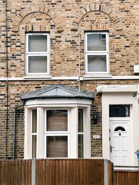Photograph of the exterior of a residential brick building with two white-framed, double-glazed sash windows on the upper floor, both with closed blinds. The ground floor features a white bay window with a triangular metal roof and a large glass pane, also with a closed blind. To the right of the bay window is a white front door with a small semi-circular window at the top and a black handle, set within a white doorframe. The building's brick wall has decorative arch accents above the upper windows, and black drainage pipes run vertically along the façade. A wooden fence partially obscures the lower part of the building, and a small white pipe runs horizontally along the exterior. The overall scene is well-lit with natural daylight, emphasizing the clean and maintained appearance of the building's surface. This setting is relevant to domestic cleaning services, such as those offered by Oven Cleaning Haringey, which specializes in surface cleaning, deep cleaning, and sanitisation of residential properties.