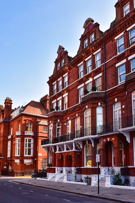 A row of red brick residential buildings with ornate architectural details, including large bay windows, decorative cornices, and wrought iron balconies, situated along a quiet street under a clear blue sky. The ground level features white steps and railings, with the entrance doors partially visible. The scene highlights the exterior condition of the buildings, emphasizing clean facades and well-maintained surfaces, which are indicative of professional exterior cleaning and maintenance services offered by Oven Cleaning Haringey, as featured on the Crouch End end of tenancy cleaning page.