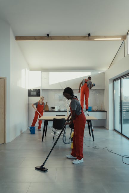 A team of three cleaning professionals in red overalls are performing cleaning tasks in a modern, well-lit kitchen with large sliding glass doors. One cleaner is using a floor vacuum cleaner on a ligh