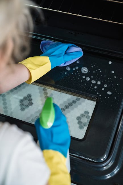 A young man with short, dark hair, wearing a light-colored hoodie with a grey vest, is opening a black, built-in electric oven in a modern kitchen. The oven is installed among white cabinetry and has