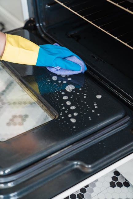 Close-up of a person's gloved hand cleaning the black glass surface of an oven door with a blue cloth, showing soap suds and streaks, in a modern kitchen with stainless steel oven interior visible. The surrounding area includes a section of a hexagonal black and white tiled floor, and the oven's metal frame is clean and reflective. The image emphasizes surface cleaning and deep cleaning efforts by Oven Cleaning Haringey, demonstrating hygienic maintenance of kitchen appliances.