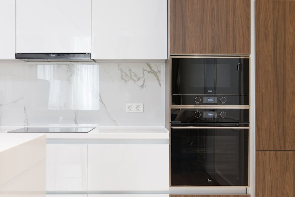 A modern kitchen featuring a white marble countertop and backsplash with subtle grey veining, adjacent to a wall with two built-in black glass ovens with stainless steel accents. The cabinetry includes sleek white upper cabinets and warm wooden panels on the right side. The kitchen surface is clean and polished, reflecting light from a nearby window, and alongside the wall, there is an electrical outlet. Visible kitchen tools include a smooth cooktop on the countertop, and the area appears to be well-maintained, consistent with surface cleaning and deep cleaning practices offered by Oven Cleaning Haringey in their domestic cleaning services.
