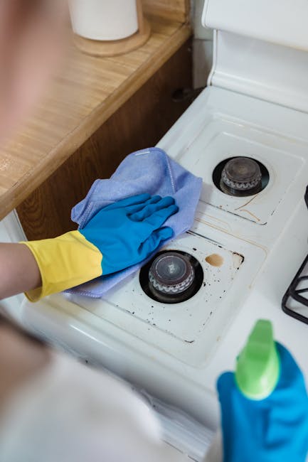 Close-up of a person's gloved hand cleaning the black glass surface of an oven door with a blue cloth, showing soap suds and streaks, in a modern kitchen with stainless steel oven interior visible. The surrounding area includes a section of a hexagonal black and white tiled floor, and the oven's metal frame is clean and reflective. The image emphasizes surface cleaning and deep cleaning efforts by Oven Cleaning Haringey, demonstrating hygienic maintenance of kitchen appliances.